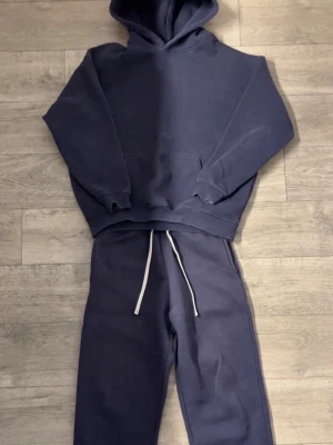 Marinblå tracksuit - Säljer ett mörkblått mjukisset med hoodie och matchande byxor. Hoodien har en stor ficka framtill och ribbade muddar. Byxorna har resår i midjan och vita dragsnören. Setet är gjort i mjukt bomullsliknande material och har en avslappnad passform.