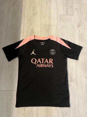 PSG T-shirt S - Snygg svart PSG matchtröja från Jordan med rosa detaljer på axlar och krage. Tröjan har tryck med Qatar Airways på bröstet och PSG-logga samt Jumpman-logga. Perfekt för fotbollsträning eller att bära till vardags.