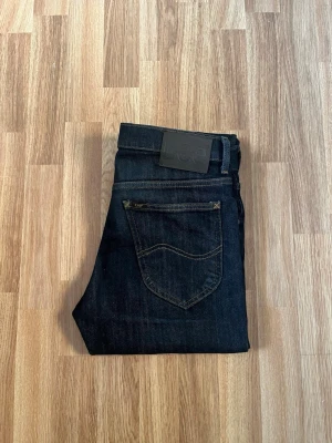 Mörkblå Lee jeans  - Snygga mörkblå jeans från Lee med klassisk femficksdesign och raka ben. Jeansen har en stilren look med mörka sömmar och ett svart Lee-läderpatch bak i midjan. Perfekta för dig som gillar en enkel och tidlös stil. Storlek W28 L32