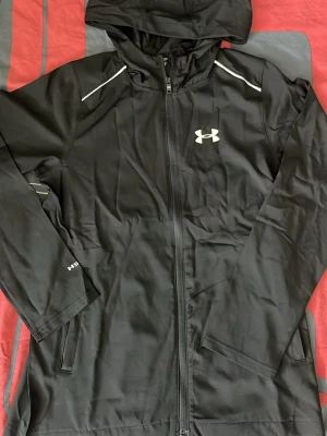 Svart hoodie från Under Armour - Snygg svart hoodie från Under Armour med dragkedja framtill och reflekterande detaljer på axlarna. Hoodien har en huva, långa ärmar och två sidofickor. Perfekt för träning eller chill. Materialet är mjukt och stretchigt för extra komfort.