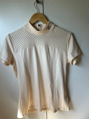 Beige polotopp (ridkläder) - Snygg beige polotopp med korta ärmar och prickigt nät över axlar och bröst. Toppen har en liten dragkedja bak i nacken och en tight passform. Perfekt för dig som gillar stilrena detaljer och sportig vibe.