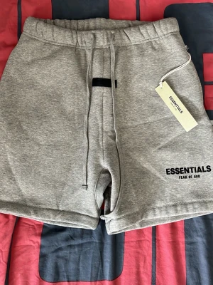Grå Essentials Fear of God shorts - Snygga grå shorts från Essentials Fear of God med broderad logga på benet. De har snörning i midjan och en relaxed passform. Perfekta för chill dagar eller streetwear-stil. Mjukt material som känns skönt mot huden.