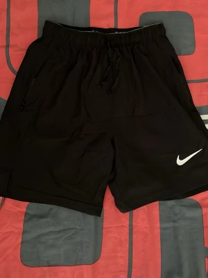 Svarta träningsshorts från Nike - Snygga svarta träningsshorts från Nike med vit logga på vänster ben. Shortsen har elastisk midja med snörning och är tillverkade i ett lätt och snabbtorkande syntetmaterial. Perfekta för gymmet eller löprundan.