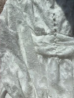 Vit spetsklänning med knappar - Romantisk vit kortklänning i spets med långärmad design och dekorativa knappar framtill. Klänningen har markerad midja och volangdetaljer i kjolen. Perfekt för sommarens alla tillfällen och ger en elegant känsla.