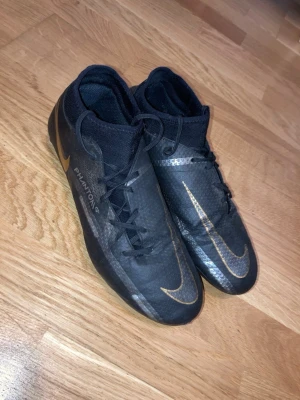 Nike Phantom GT Fotbollsskor - Säljer ett par använda svarta mid-sock Nike Phantom GT fotbollsskor med gulddetaljer.Skorna har svarta snören och en svart sula. Skorna är använda ett par gånger, men funkar lika bra att använda flera ggr.