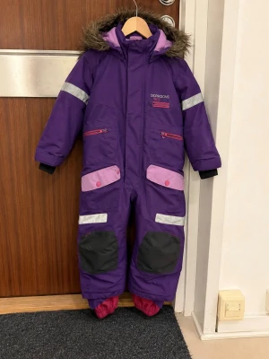 Täcköverall / Overall från Didriksons - Didriksons vinteroverall i storlek 80. Overall i lila med rosa och svarta detaljer från kvalitetsmärket Didriksons. Utrustad med luva med avtagbar fuskpäls, dragkedja framtill, reflexdetaljer samt förstärkta partier på knän och bakdel för ökad slitstyrka. Ärmslut och benslut är justerbara för bra passform.  Nypris för motsvarande Didriksons vinteroverall ligger idag omkring 1 500 kr i butik.  Mycket gott skick, se bilder