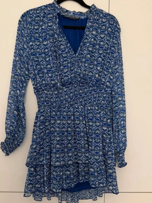 Blå mönstrad kortklänning - Säljer en blå kortklänning med vitt mönster och långärmad design. Klänningen har volangdetaljer vid kjolen, resår i midjan och vid ärmsluten samt en lätt v-ringad omlott upptill. Perfekt för dig som gillar färg och mönster!