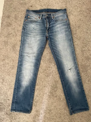 Ljusblå Levi’s jeans med slitning - Säljer ett par ljusblå jeans med raka ben och snygga slitningar på framsidan. Jeansen har klassisk femficksdesign, knappgylf och kontrastsömmar. Materialet är denim i bomull och passformen är avslappnad men ändå stilren.