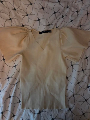 Beige topp med puffärm från Vero Moda - Trendig beige topp från Vero Moda med breda puffärmar och v-ringad hals. Toppen är stickad i mjukt material och har en slim passform som framhäver siluetten. Perfekt för dig som vill ha en stilren men ändå unik look.