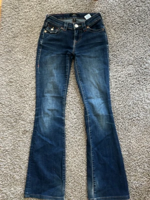 Blå bootcut jeans med fickdetaljer - Säljer ett par blå bootcut jeans med snygga slitningar och utställda ben. Jeansen har coola fickdetaljer bak med kontrastsömmar och knappar. Perfekta för dig som gillar en retro vibe och vill sticka ut lite extra.