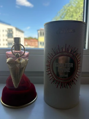 Jean Paul Gaultier Divine Couture parfym - Unik parfymflaska från Jean Paul Gaultier Divine Couture med en kroppsinspirerad design i guld och rött. Flaskan har detaljer i metall och glas, och kommer med en elegant vit förpackning med gulddetaljer. Perfekt för dig som gillar lyxiga och kreativa dofter.