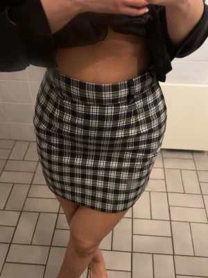 Svartvit rutig kortkjol - Snygg svartvit rutig kortkjol med hög midja och figurnära passform. Kjolen har klassiskt rutmönster och passar perfekt till en trendig vardagslook. Enkel att matcha med både crop tops och stickade tröjor. Finns även ett matchade bälte till som tillkommer