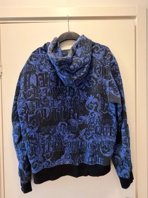 Blå hoodie från Versace Jeans Couture - Snygg blå hoodie med svart botten och all-over print i blått och svart från Versace Jeans Couture. Hoodien har dragkedja framtill, huva och ribbade muddar. Materialet är mjukt och bekvämt, perfekt för en trendig streetwear-look.