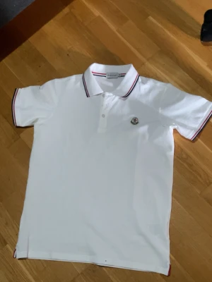 Vit Moncler pikétröja XL - Säljer en klassisk vit pikétröja från Moncler i storlek XL. Tröjan har korta ärmar, krage med röd och marinblå rand samt Moncler-logga på bröstet. Perfekt till en clean och stilren look.