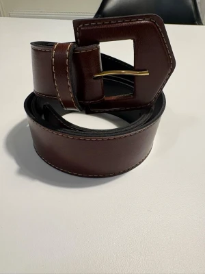 Brett brunt skärp med fyrkantigt spänne - Stilrent och brett brunt skärp i skinn med markerade vita sömmar och ett stort fyrkantigt spänne i guldtonad metall. Perfekt accessoar för att lyfta din outfit med en klassisk touch.