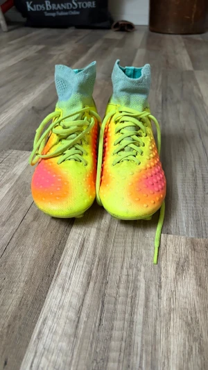 Nike gula och orange fotbollsskor - Säljer ett par grymma fotbollsskor från Nike i neon-gult och orange med svart swoosh. Skorna har hög strumpkrage i ljusblått och knottrig ovansida för bättre bollkontroll. Perfekta för dig som vill sticka ut på planen!