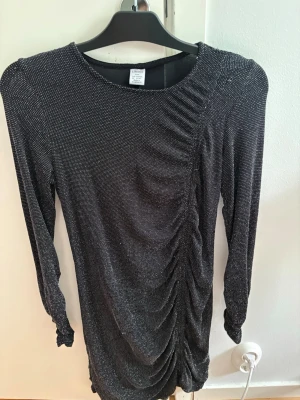 Svart glittrig klänning från Lindex - Snygg svart klänning från Lindex med glittrigt tyg och rynkade detaljer framtill. Klänningen har lång ärm och en kroppsnära passform, perfekt för dig som vill sticka ut lite extra.