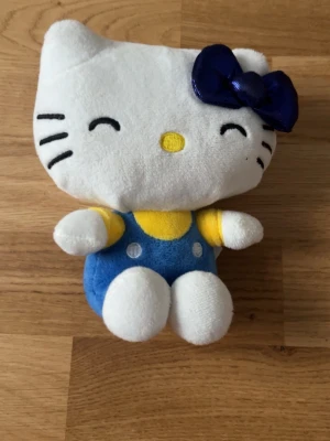 Hello Kitty mjukisdjur med blå rosett - Söt Hello Kitty mjukisdjur i vitt plyschmaterial med blå och gul klänning samt en stor blå glansig rosett på huvudet. Ansiktet har broderade detaljer och figuren har en klassisk design med rund form och små ben.