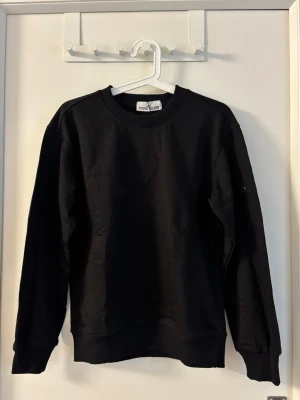 Svart sweatshirt från Stone Island - Snygg svart sweatshirt från Stone Island med rund halsringning och ribbade muddar. Tröjan har en diskret logoknapp på ärmen och är tillverkad i mjukt bomullsmaterial. Perfekt för en clean och stilren look.