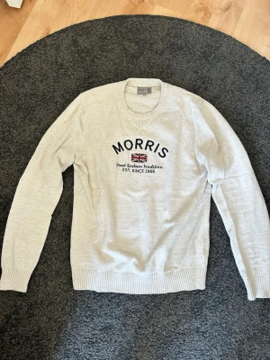Stickad tröja från Morris - Vit/krämvit stickad tröja från Morris med rund halsringning och broderad text samt brittisk flagga på bröstet. Tröjan har långa ärmar och ribbade muddar vid ärmslut och nederkant. Perfekt för en stilren och avslappnad look.