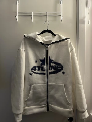 Vit hoodie med 4TUNE-tryck - Säljer en vit hoodie med dragkedja och stor huva. På framsidan finns ett blått 4TUNE-tryck med stjärnor. Hoodien har två stora fickor framtill och är gjord i ett mjukt, fleece-liknande material. Perfekt för en chill och avslappnad stil.