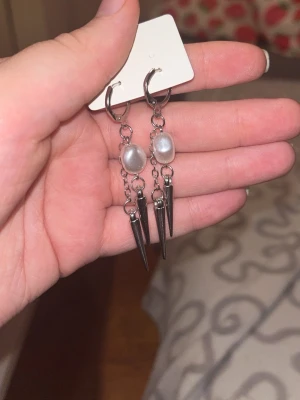 Silvriga örhängen med pärlor och spikes - Säljer ett par trendiga silvriga örhängen med kedjor, vita pärlor och svarta spikes. Örhängena har en cool och edgy design som sticker ut. Perfekta för dig som vill ha något unikt och snyggt till din stil.