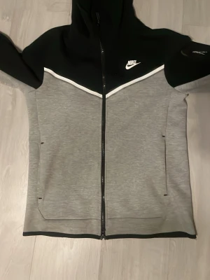 Nike tech fleece  - Snygg hoodie från Nike i grått och svart med vit detalj över bröstet. Hel dragkedja framtill, två fickor och klassisk Nike-logga på bröstet. Tillverkad i mjukt material och har huva för extra stil och komfort. (Nike tech)