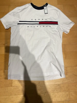 Vit Tommy Hilfiger t-shirt med tryck - Vit t-shirt från Tommy Hilfiger med svart ribbad krage och ett klassiskt tryck med logga och ränder i svart, vitt och rött över bröstet. T-shirten har korta ärmar och är gjord i mjuk bomull för en skön känsla.
