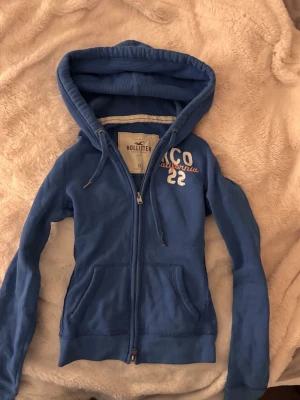 Blå zip-hoodie vintage Hollister - Snygg blå hoodie från Hollister med dragkedja, huva och fickor framtill. Broderad text 'A&CO California 22' på bröstet. Perfekt för en avslappnad och cool stil. Mjuk bomullsblandning och ribbade muddar vid ärmslut och nederkant.
