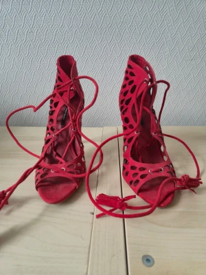 Röda pumps med snörning och hålmönster - Snygga röda pumps med öppen tå, höga klackar och dekorativt hålmönster i mockaimitation. Skorna har snörning framtill som kan knytas runt vristen för en cool look. Perfekta för dig som vill sticka ut med ett par statement-heels.