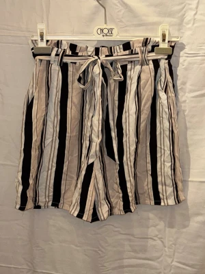 Randiga shorts med knytband - Snygga shorts med vertikala ränder i svart, beige och vitt. De har ett matchande knytband i midjan och avslappnad passform. Perfekta för varma dagar och ger en trendig look.