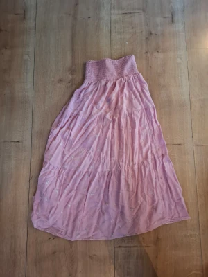 Ljusrosa långkjol med smock - Söt ljusrosa långkjol med smock i midjan och volangdetaljer. Kjolen har ett tunt, flowy tyg och små broderade detaljer som ger en romantisk känsla. Perfekt för dig som gillar en feminin och avslappnad stil.
