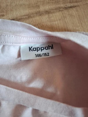 Ljusrosa t-shirt från Kappahl - En enkel och stilren ljusrosa t-shirt från Kappahl. T-shirten har korta ärmar och är gjord i mjuk bomull, perfekt för en avslappnad look. Passar till det mesta och är lätt att matcha med andra plagg. Den är tajt i midjan. 