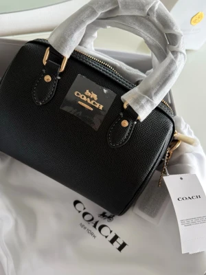 MINI BAG - Coach handväska i svart med dubbla handtag och snygga gulddetaljer. En stilren liten modell med klassisk form, i modellen Mini Rowan Black Bag. All tillbehör medföljer med, i äkta läder.
