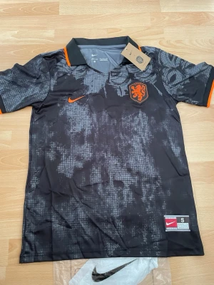 Svart Nederländerna matchtröja Nike - Säljer en svart matchtröja från Nike för Nederländernas landslag. Tröjan har orange detaljer, krage och ärmslut samt ett orange lejonmärke på bröstet. Materialet är lätt och andas, perfekt för fotboll eller träning. Snyggt mönster över hela tröjan.