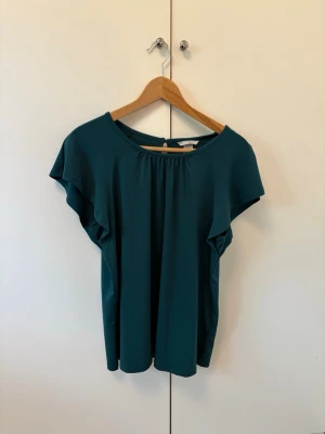 Mörkgrön blus med kort ärm - Snygg mörkgrön blus med rund halsringning och korta, löst sittande ärmar. Blusen har ett enkelt men elegant fall och en liten knappdetalj i nacken. Perfekt att styla med jeans eller kjol.