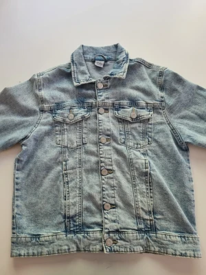 Ljusblå klassisk jeansjacka - Snygg ljusblå jeansjacka med raka linjer och klassisk krage. Jackan har två bröstfickor med lock och silverfärgade knappar framtill. Perfekt att slänga över en hoodie eller t-shirt för en avslappnad look.