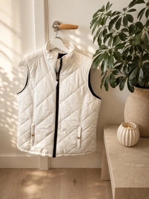 Vit quiltad dunväst med dragkedja - Stilren vit dunväst med hög krage och svart dragkedja framtill. Västen har två fickor med knappstängning och ett quiltat mönster som ger en modern look. Perfekt att slänga över en hoodie eller tröja för extra värme. Materialet är lätt och smidigt.