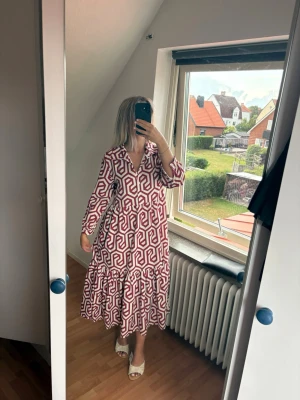 Vit och röd mönstrad långklänning - Säljer en långklänning med unikt rött och vitt geometriskt mönster. Klänningen har v-ringning, långa ärmar och volang nedtill. Perfekt för dig som gillar statement-plagg med skön passform och trendig look.