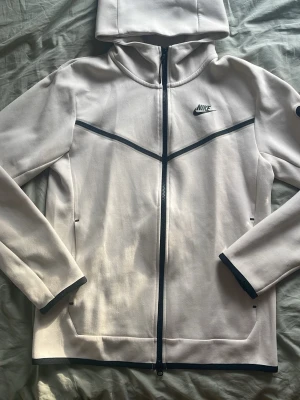 Off-white Nike Tech Fleece - Snygg ljusgrå hoodie från Nike med hel dragkedja och svarta detaljer. Har huva, två fickor och diskret Nike-logga på bröstet. Materialet känns mjukt och sportigt, perfekt för chill eller träning. Svarta linjer ger en modern look.