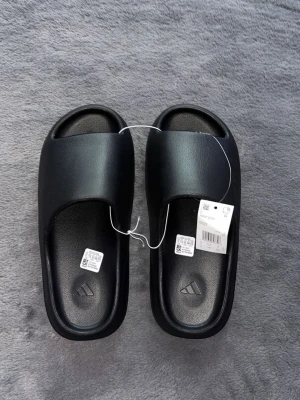 Svarta Yeezy Slide tofflor från Adidas - Säljer ett par svarta Yeezy Slide tofflor från Adidas. De har en clean och modern design med bred rem över foten och räfflad sula för extra grepp. Tillverkade i ett mjukt och bekvämt syntetmaterial som passar perfekt för chill dagar hemma eller ute.