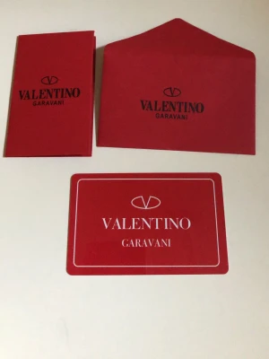 Valentino Garavani kort och kuvert - Säljer ett set med rött Valentino Garavani-kort, matchande kuvert och liten folder. Alla delar är i papper med tydlig logga och text i svart och vitt. Perfekt till present eller samling.