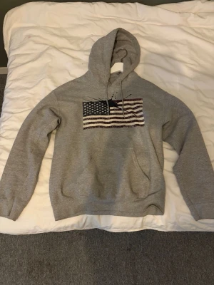 Grå hoodie med amerikansk flagga - Säljer en grå hoodie med huva och dragsnören. På bröstet finns ett tryck av den amerikanska flaggan i slitad stil. Hoodien har en klassisk känguruficka framtill och ribbade muddar vid ärmar och nederkant. Perfekt för en chill och avslappnad look.