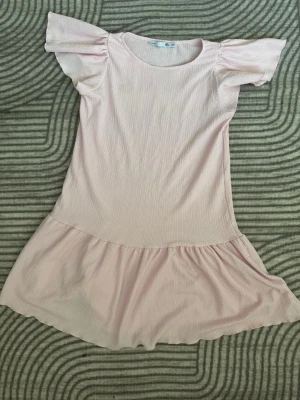 Ny Ljusrosa volangklänning med puffärm  - Söt ljusrosa kortklänning med volang nertill och puffiga kortärmar. Klänningen har rund halsringning och är gjord i ett mjukt, ribbat material som ger en bekväm känsla. Perfekt för dig som gillar en feminin och avslappnad stil.