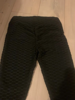 Svarta scrunch-leggings med bikakemönster - Säljer ett par svarta leggings med scrunch-effekt och bikakemönstrad struktur. De är stretchiga och har hög midja, perfekta för en trendig och bekväm look. Materialet känns mjukt och följsamt mot kroppen.
