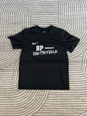 Svart Nike BP Knatteskola t-shirt - Svart t-shirt från Nike med tryck 'BP Knatteskola' och 'Stadium' på bröstet. Klassisk sportmodell med rund hals och korta ärmar. Tillverkad i ett lätt och funktionellt material som passar perfekt till träning eller fotboll.