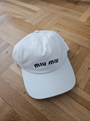 Vit keps från Miu Miu - Snygg vit keps från Miu Miu med svart broderad logga framtill. Klassisk böjd skärm och justerbar baksida. Tillverkad i bomull för en bekväm och luftig känsla. Perfekt accessoar för en trendig och avslappnad stil.