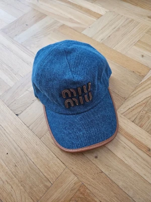 Blå denimkeps från Miu Miu - Snygg blå keps i denim från Miu Miu med bruna detaljer och broderad logga framtill. Skärmen har en brun kant i läderimitation som ger en extra cool touch. Perfekt accessoar för att lyfta din streetwear-look.