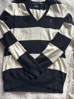 Randig tröja från Ralph Lauren - Snygg långärmad tröja från Ralph Lauren med breda svarta och vita ränder. Tröjan har V-ringning och en lila broderad logga på bröstet. Tröjan är i fint skick pga ingen användning och säljer pga att jag inte använder den. 