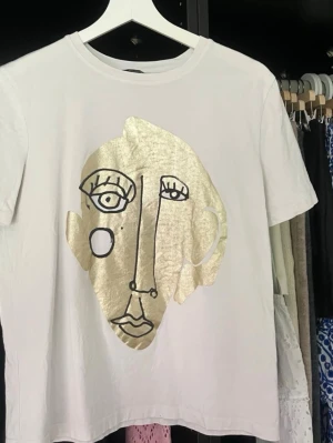 Vit t-shirt med guld ansiktsmotiv - Vit t-shirt i bomull med korta ärmar och ett stort abstrakt ansikte i guld och svart tryck på framsidan. T-shirten har en normal passform och känns riktigt trendig med sin unika design.